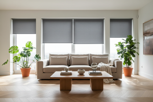Modern ve şık bir salon. Geniş pencerelerde sade ve zarif koyu gri stor perdeler düzgün şekilde takılı. Perdeler yarı açık, içeri süzülen gün ışığı ile ortam ferah ve aydınlık görünüyor. Açık renk parke zemin, bej koltuk, ahşap sehpa, birkaç dekoratif obje ve yeşil bitkiler bulunuyor. Ortam huzurlu, estetik ve davetkar olmalı. Görsel yüksek çözünürlüklü, gerçekçi ve profesyonel olmalı